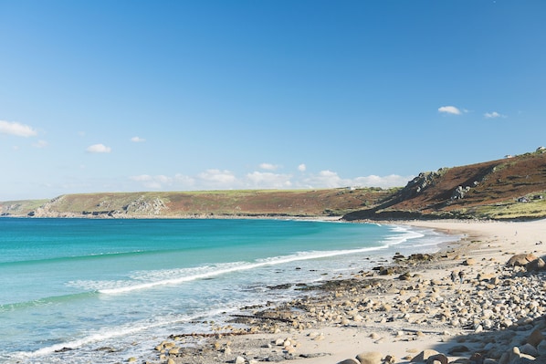 A coastal beach with rocky shoreline, clear blue water, and distant hills.