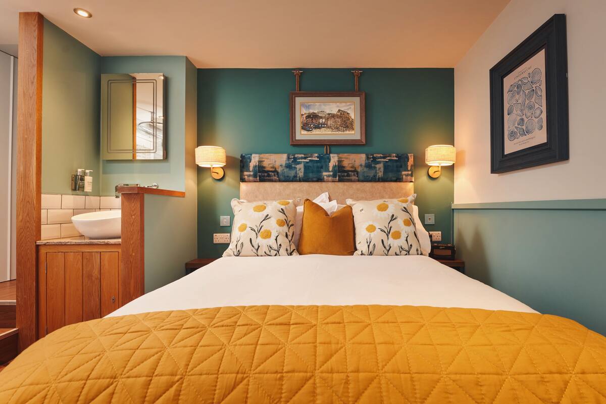A hotel room with a large bed, a wooden headboard, a wall-mounted mirror, a framed picture, and a bedside table with a lamp.