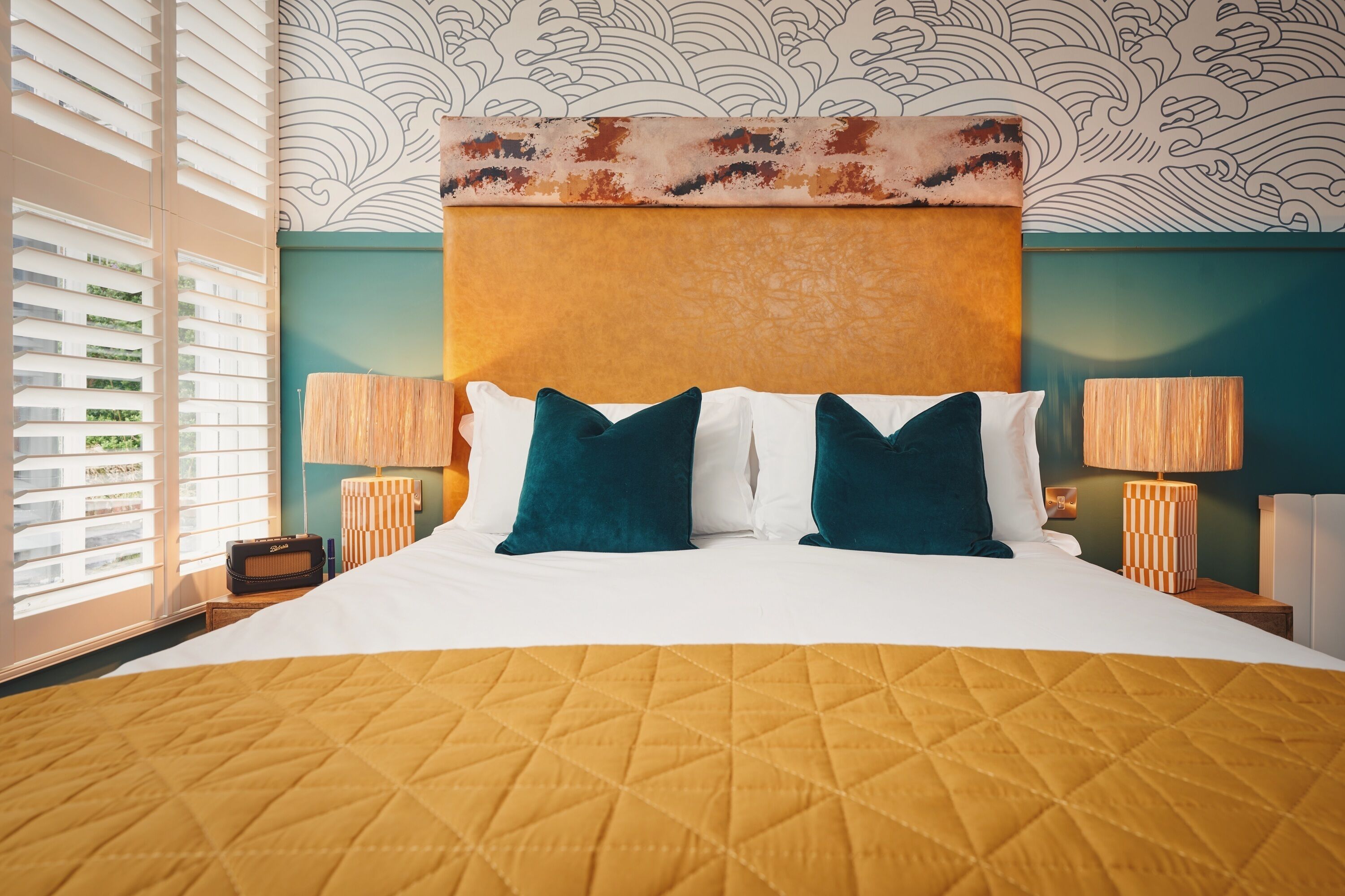 A hotel room with a large bed, two bedside lamps, a radio, and a view of greenery through the window blinds.