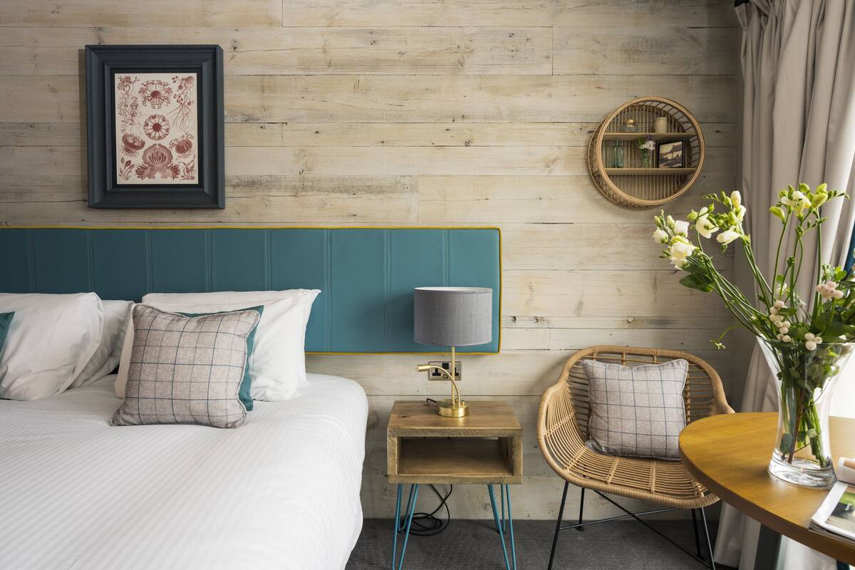 A hotel room with a bed, a bedside table with a lamp, a wicker chair, a round mirror, and a vase with flowers.