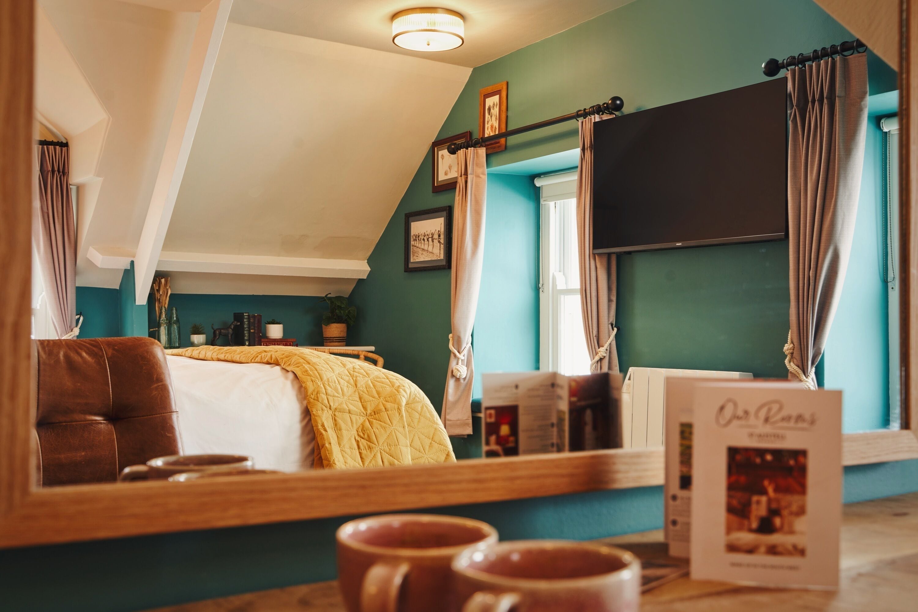 Un dormitorio con una cama, un espejo grande, una ventana con cortinas y una mesita de noche con un libro y una taza.