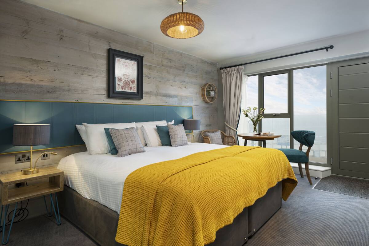 A modern hotel room with a large bed, a bedside table with a lamp, a small table with a vase, and a view of the city through large windows.