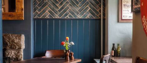 A dining area with a wooden table, chairs, and a herringbone-patterned wall. There are two bottles on the table, and a vase with flowers on the wall.