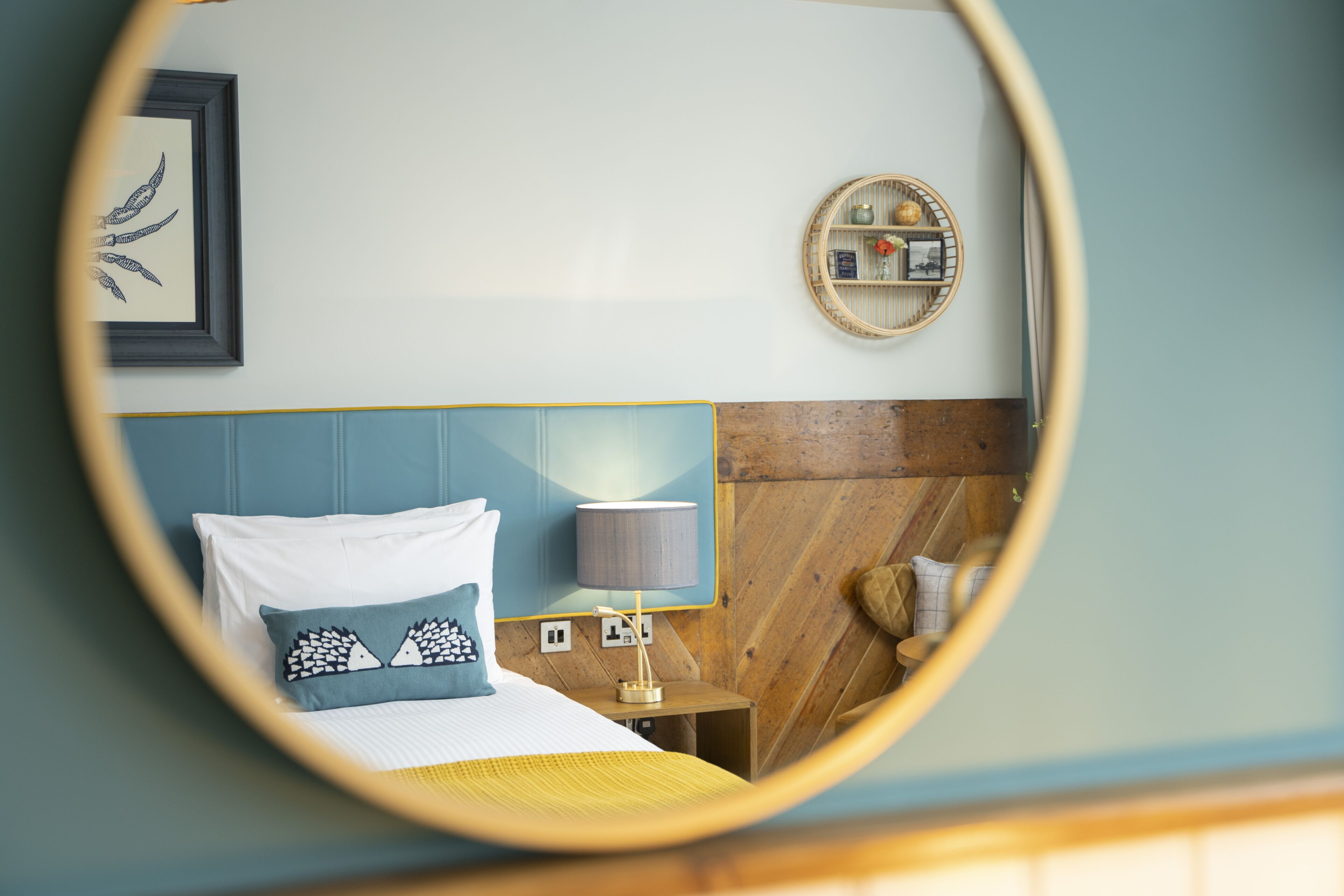 Un dormitorio con cama, mesita de noche con lámpara, espejo y estante en la pared con objetos decorativos.
