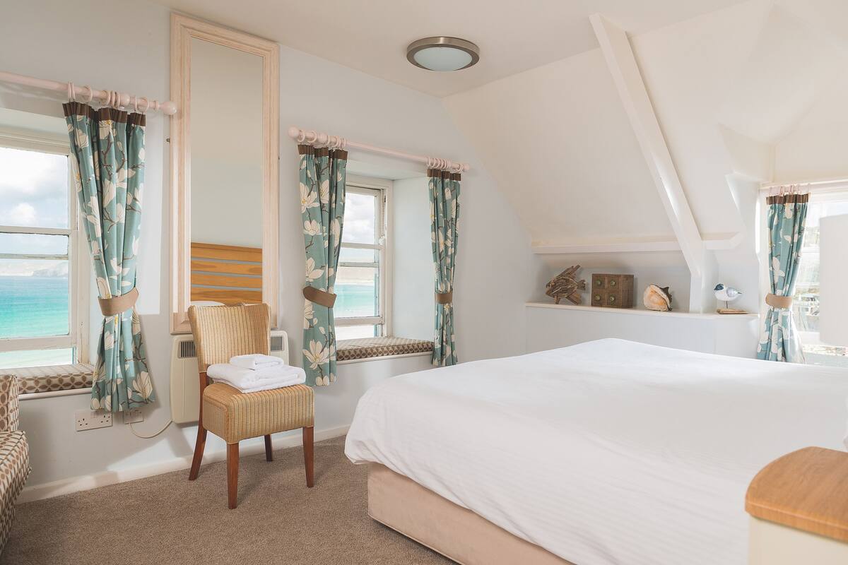 A bedroom with a large bed, a window seat with curtains, a chair, and a view of the sea.