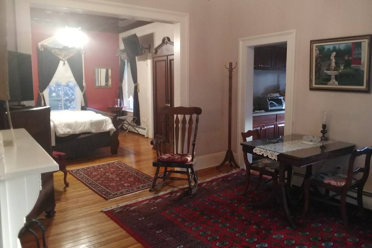 A room with a bed, a desk, a chair, a dining table, a coat rack, and a painting on the wall.