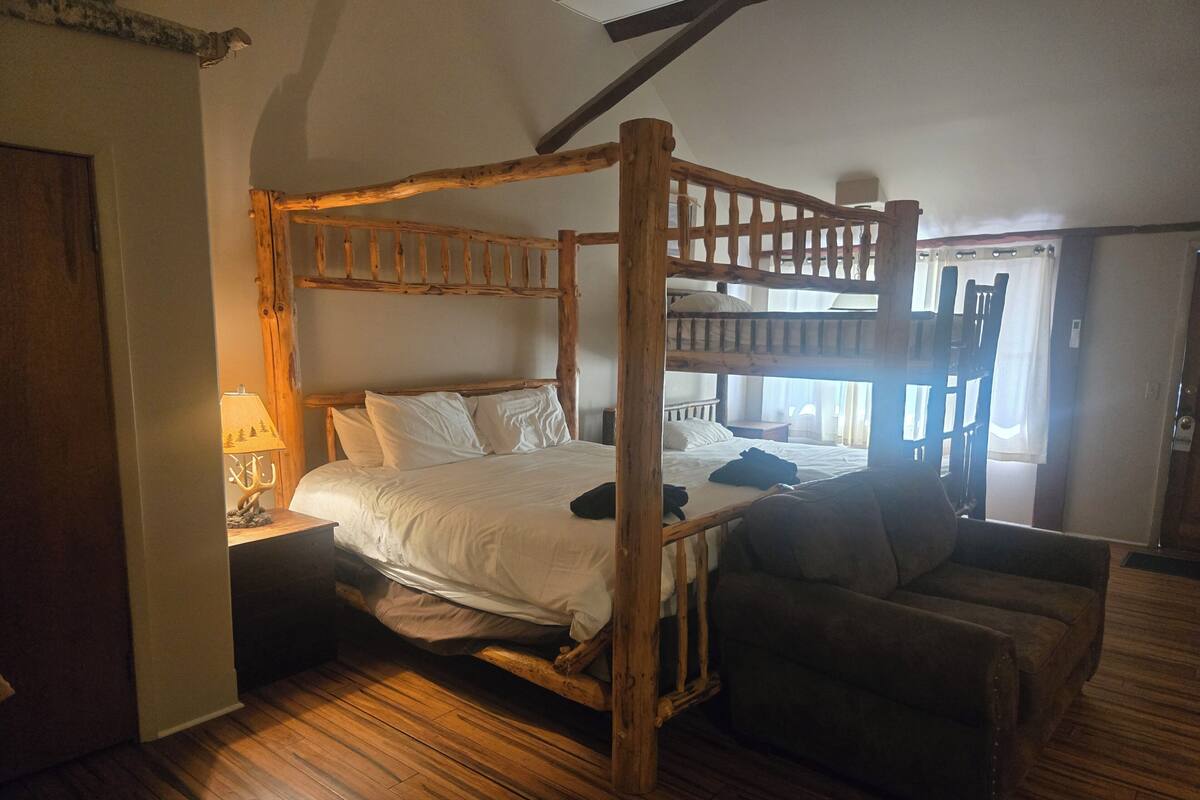 A rustic wooden bunk bed with a wooden headboard, a bedside table with a lamp, and a sofa.