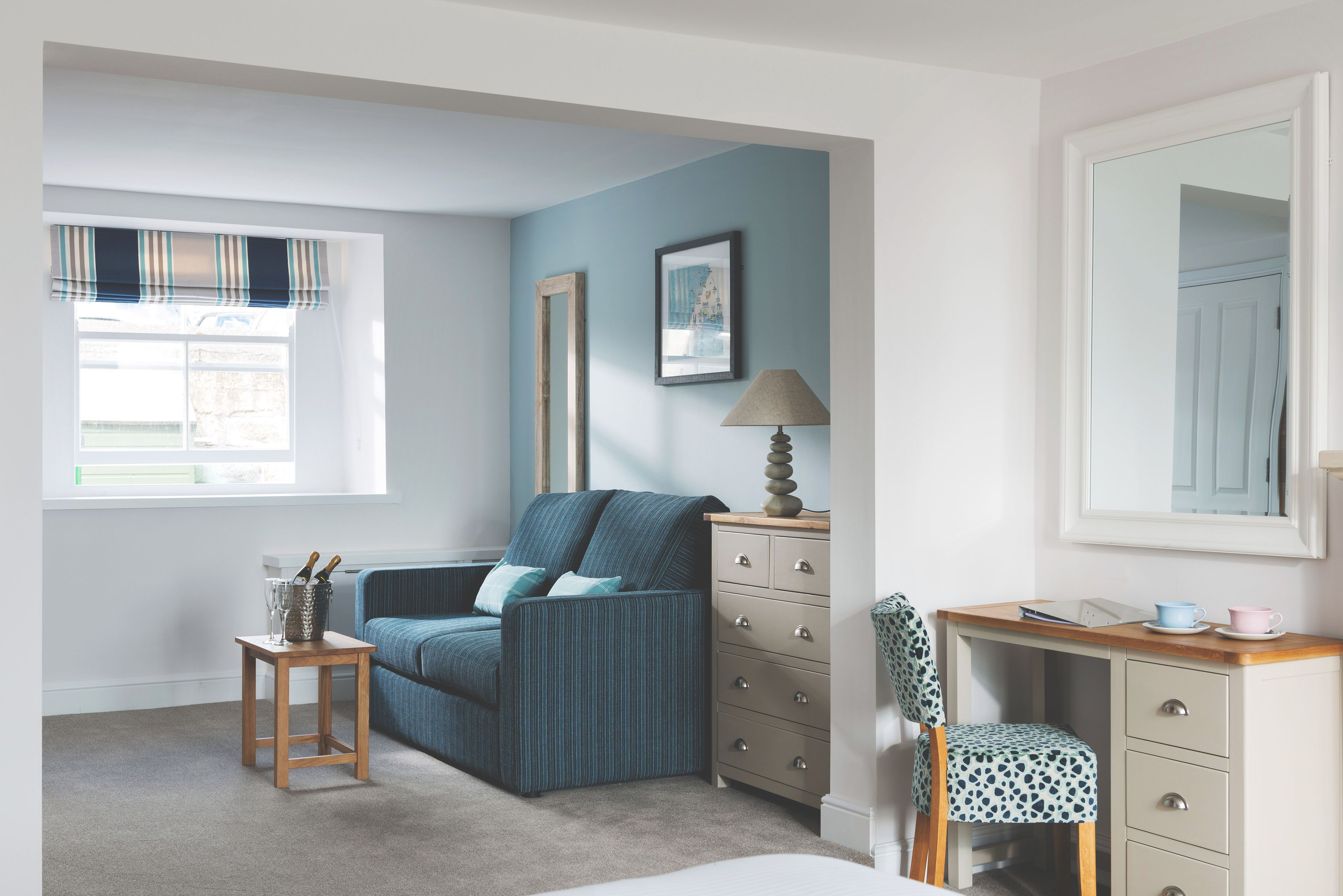 Een kamer met een blauwe bank, een houten tafel met een lamp, een dressoir en een stoel.