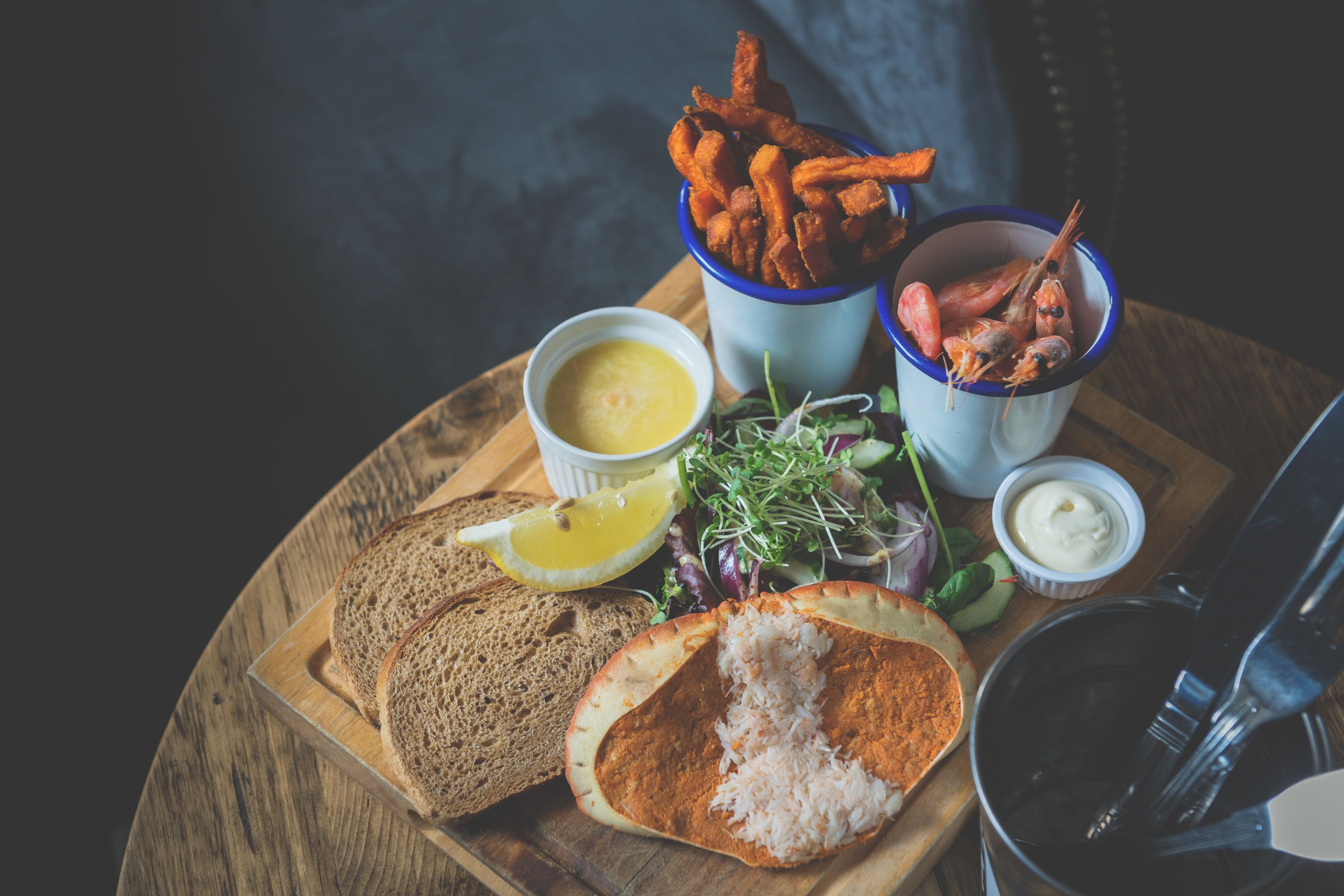 Una tabla de madera con diversos alimentos, como pan, una ensalada y una taza de camarones.