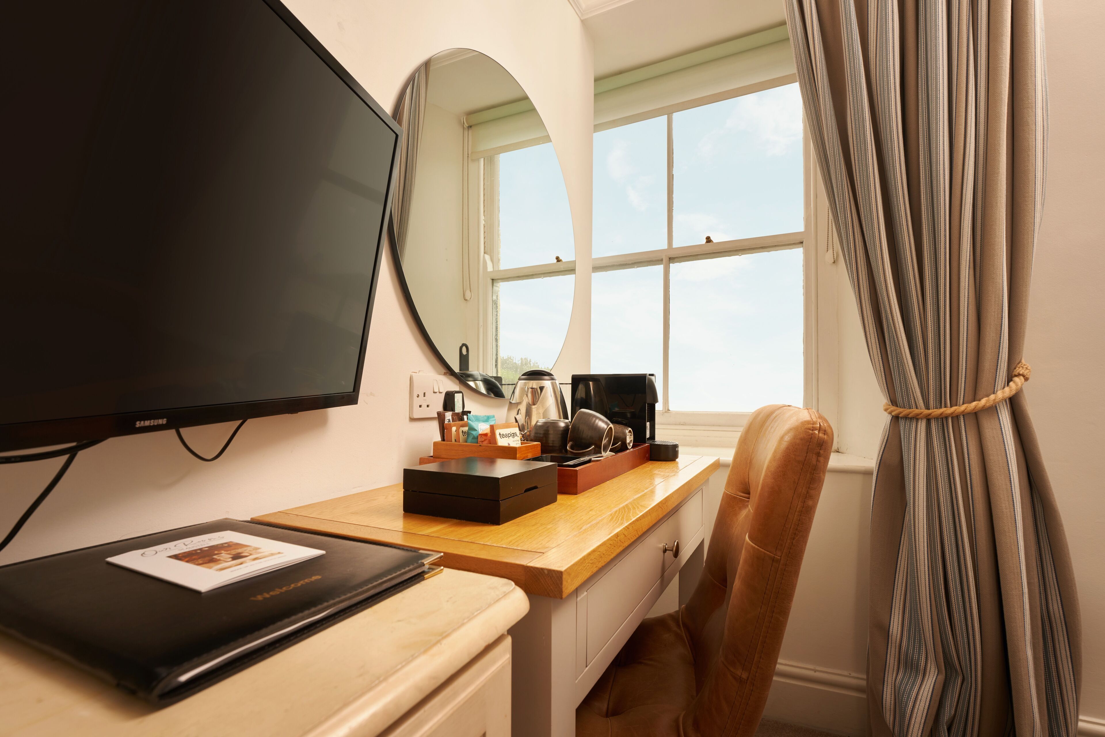 Habitación de hotel con escritorio, televisor, espejo, ventana con cortinas y una silla.