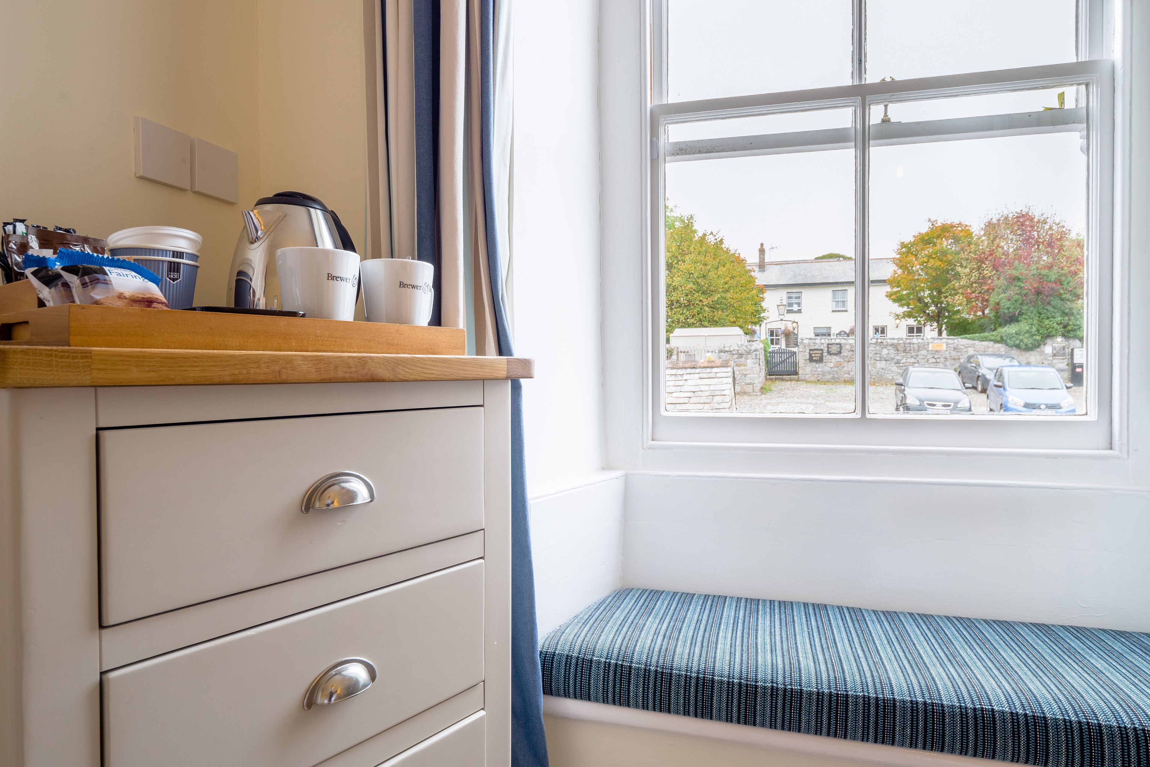 A room with a window, a bench, and a kitchenette area with a coffee maker and cups.