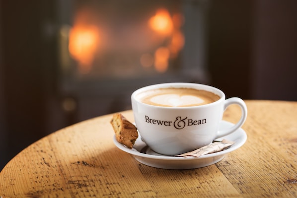 A white cup with "Brewer & Bean" logo on a wooden table with a biscuit and tea bag beside it.