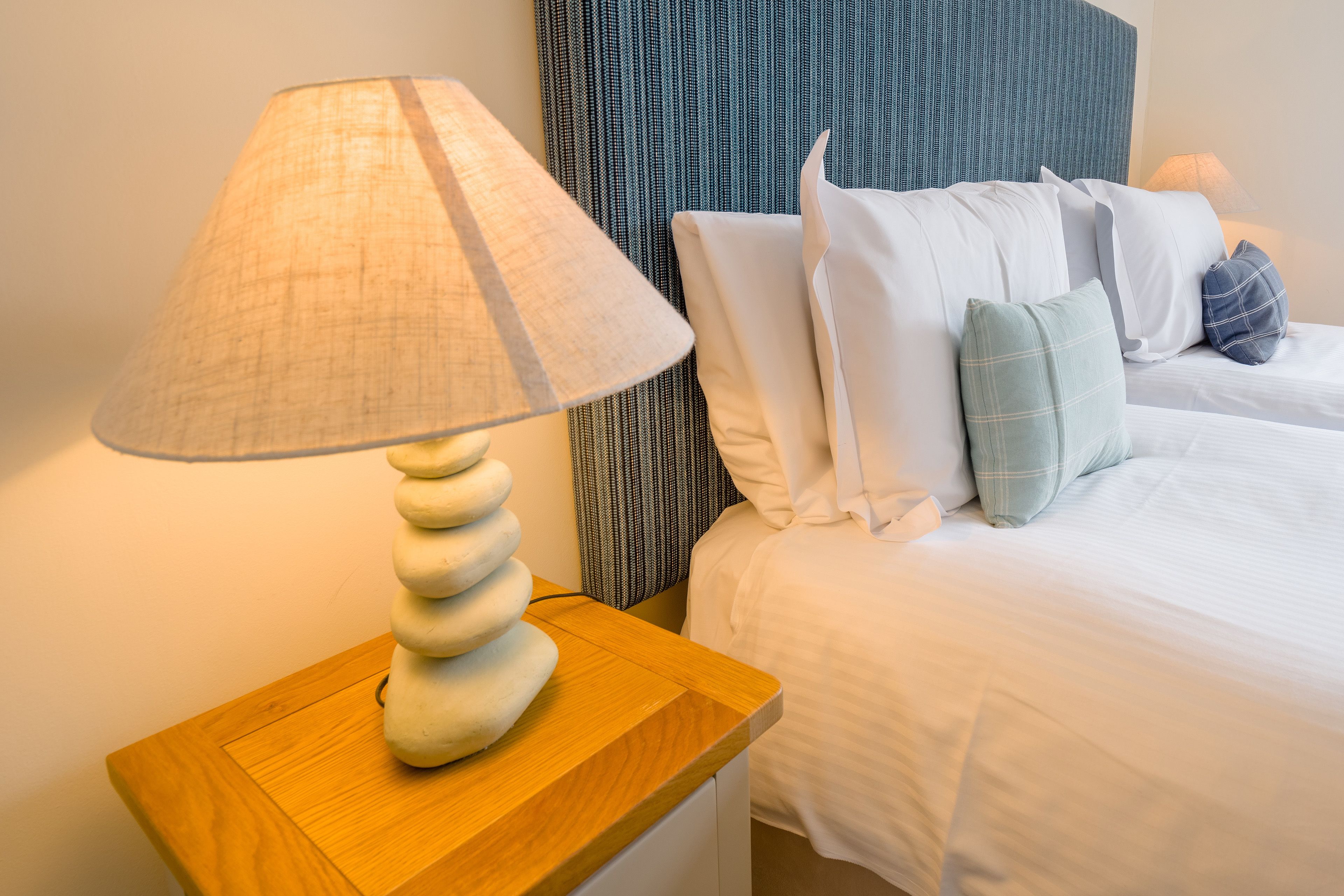A neatly made bed with white linens and multiple pillows, a wooden nightstand with a lamp, and a headboard with a striped pattern.