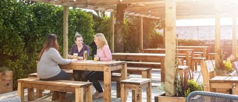 An outdoor patio with wooden tables and benches, surrounded by greenery and a pergola structure.