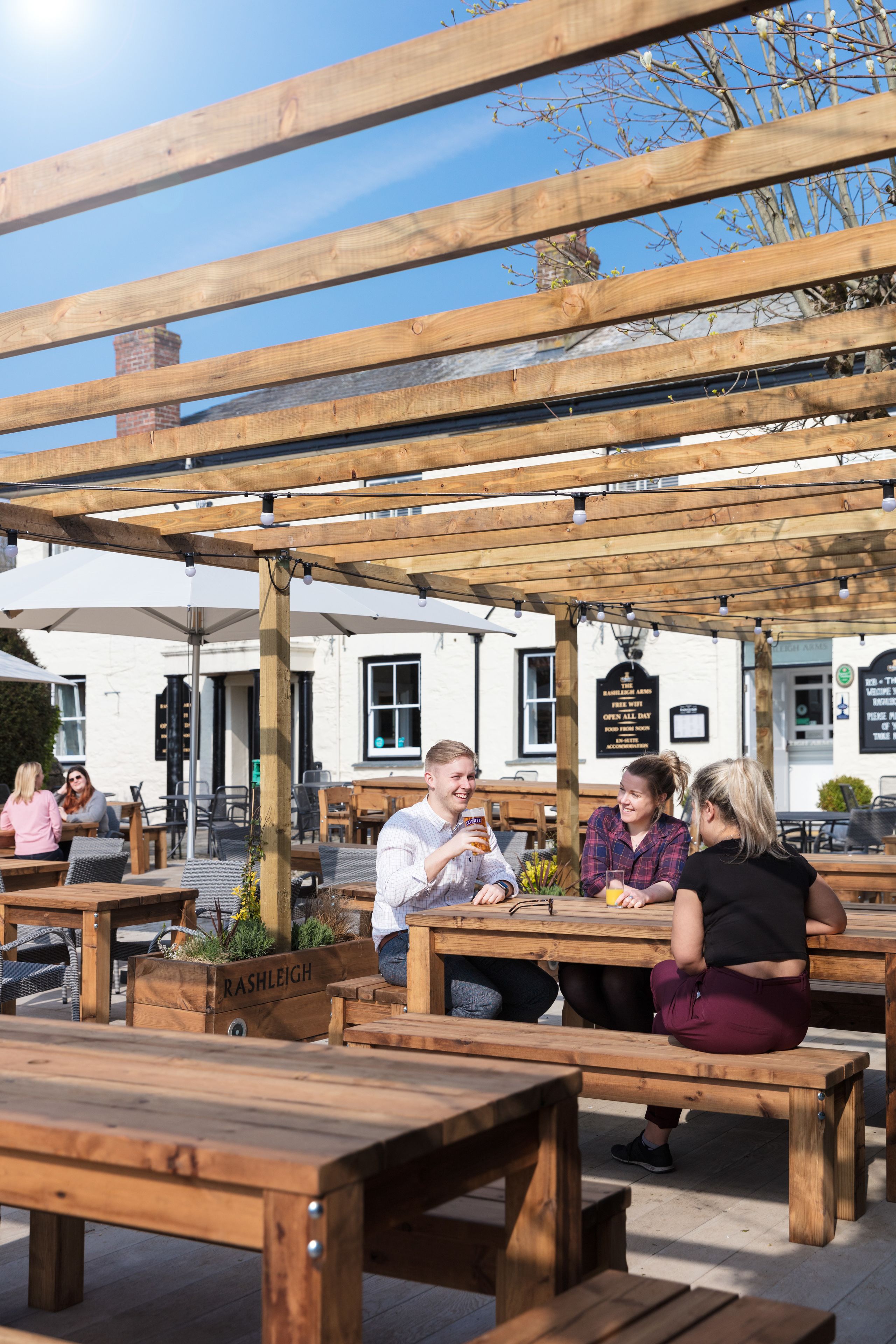An outdoor seating area with wooden benches and tables, a pergola overhead, and a building with signage in the background.