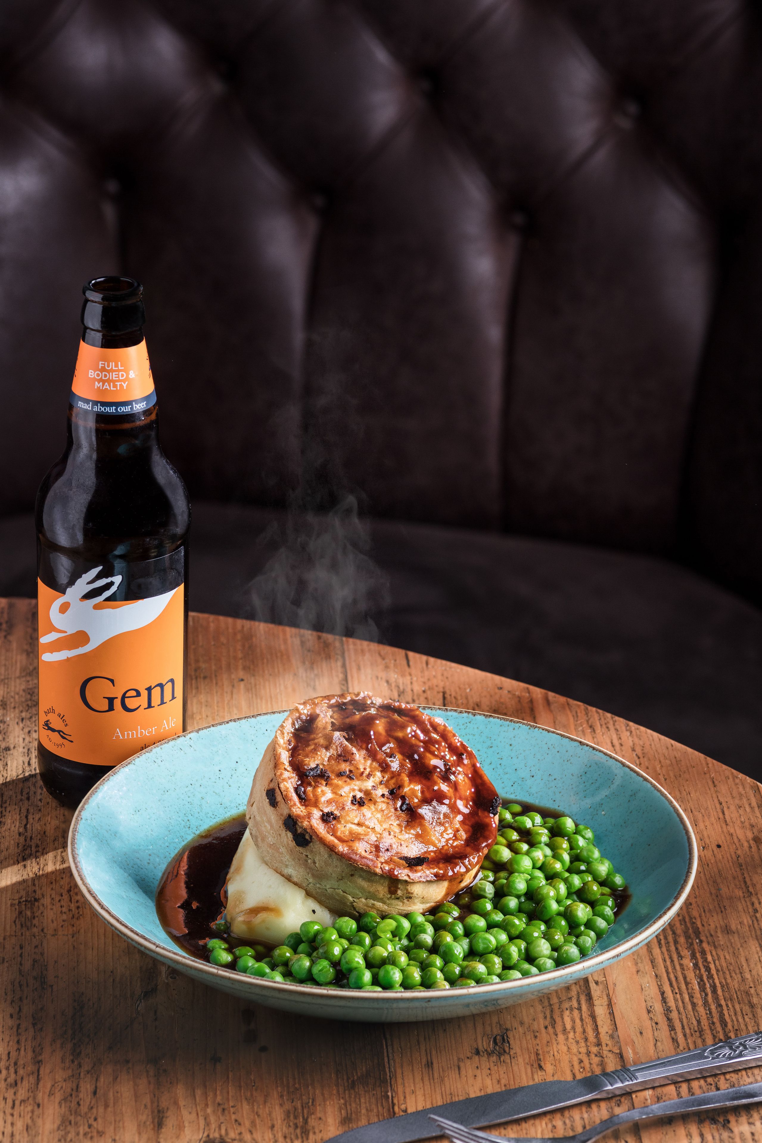 Un plato con un pastel de carne, guisantes y una botella de cerveza sobre una mesa de madera.