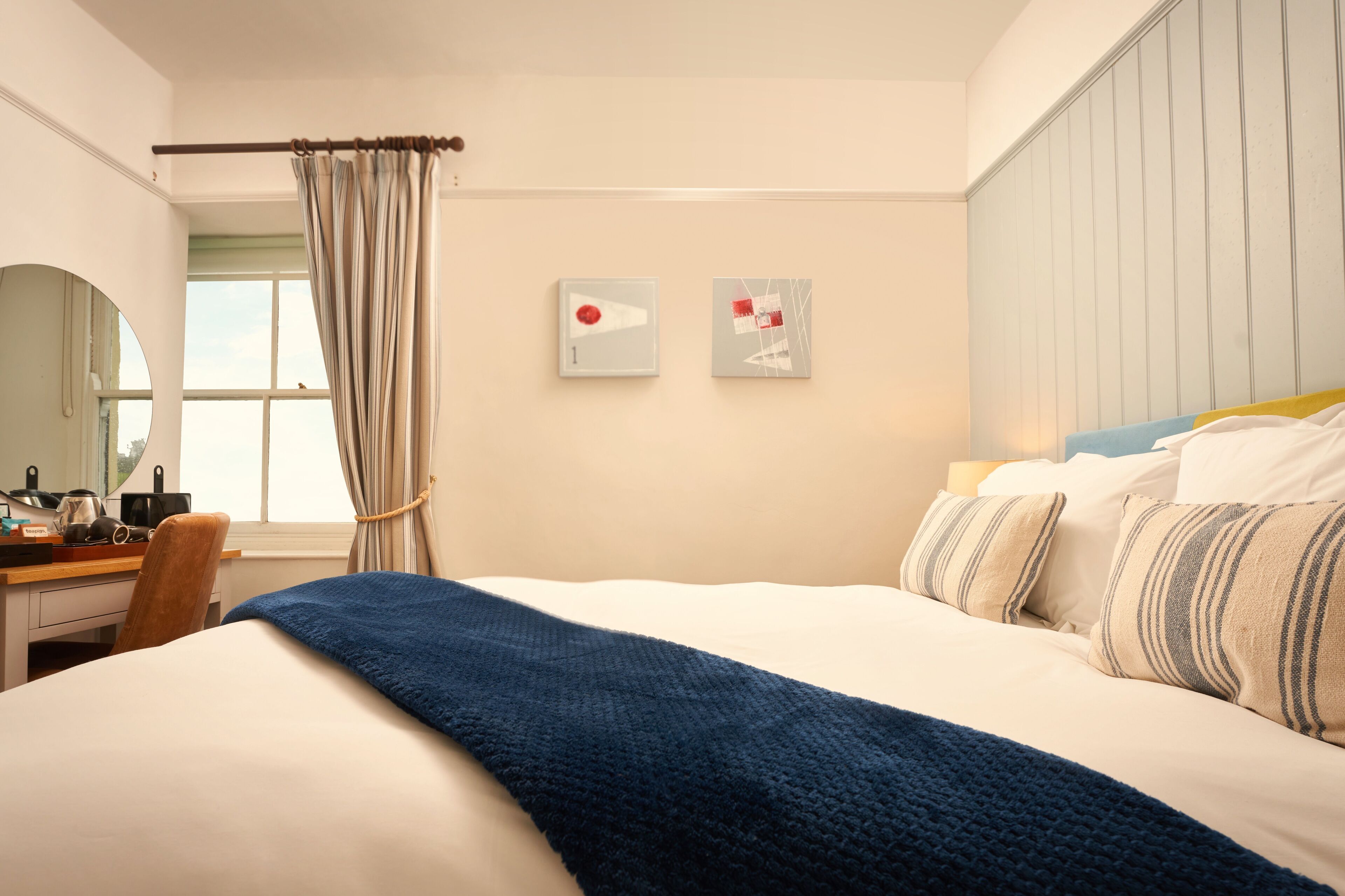 Ein Hotelzimmer mit einem Bett, einem Schreibtisch, einem Sessel, einem Fenster mit Vorhängen und zwei Wandbildern.