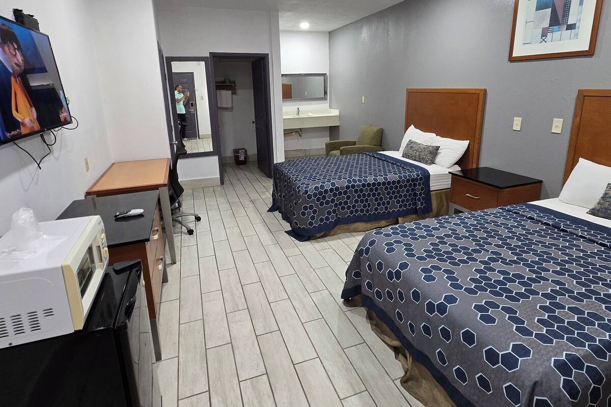 A hotel room with two beds, a desk, a television, a bathroom, and a painting on the wall.