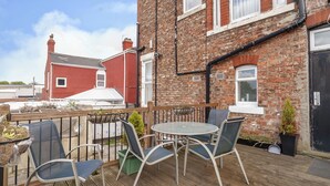 A wooden deck with a round table and chairs, a small garden area with plants, and a brick building in the background. - Kingswood (Stockton-on-Tees)