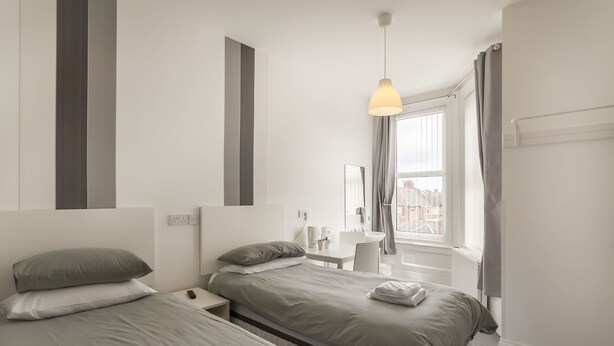 A modern hotel room with two beds, a small desk, a window with curtains, and a hanging light fixture. - Kingswood (Stockton-on-Tees)
