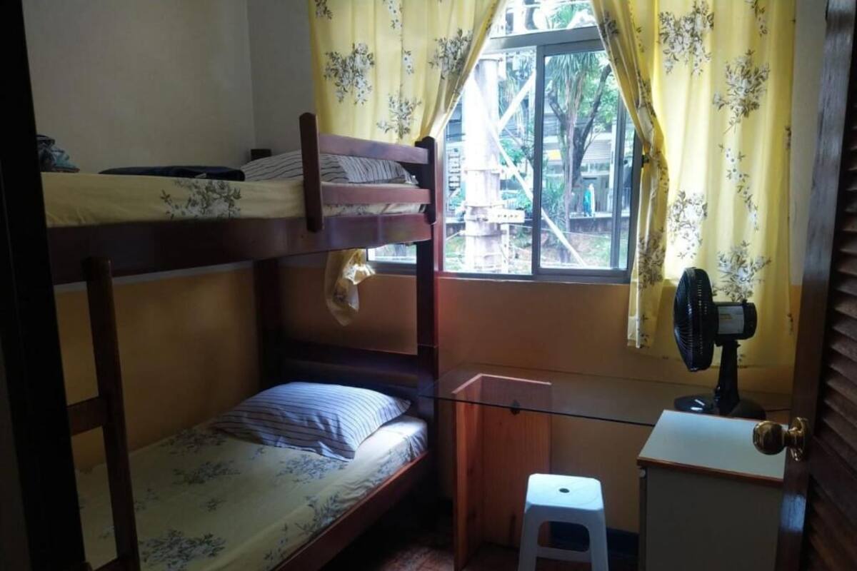 A dormitory room with two bunk beds, a small desk with a fan, a stool, and a window with curtains.