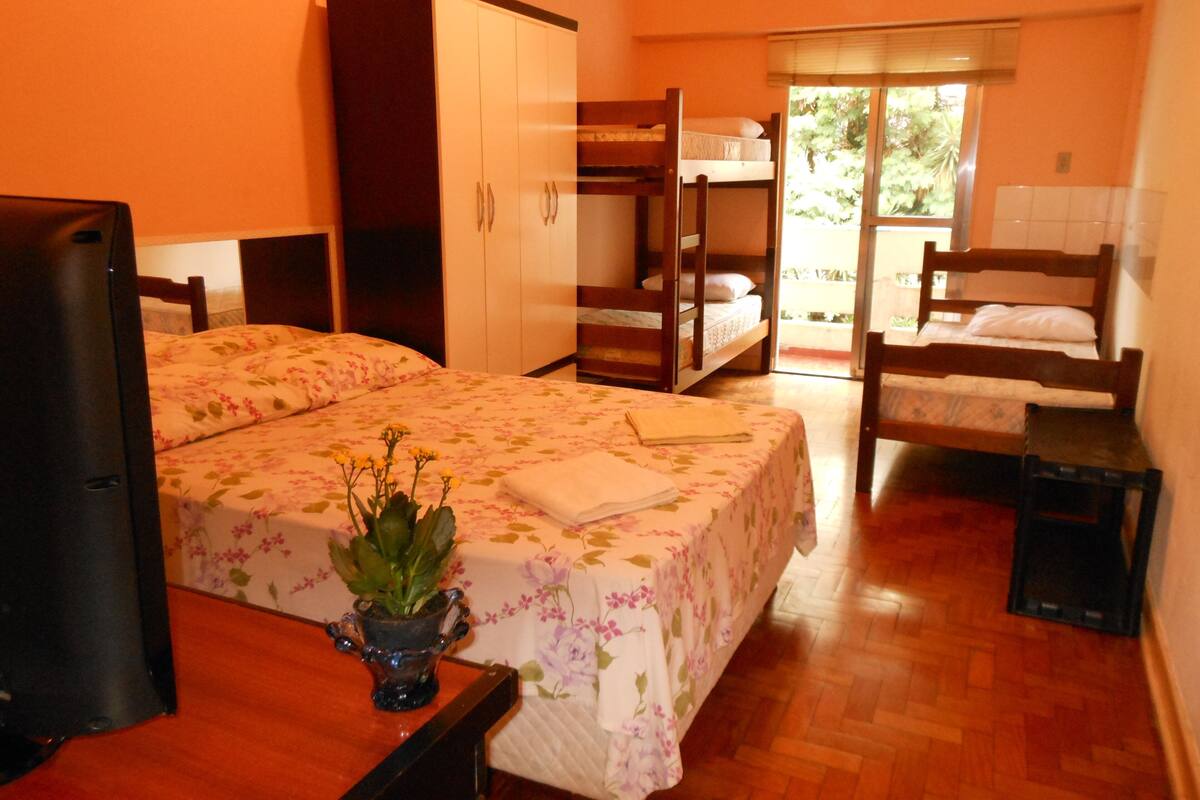 A room with a bunk bed, a desk with a computer, a wardrobe, and a window with a view of greenery.