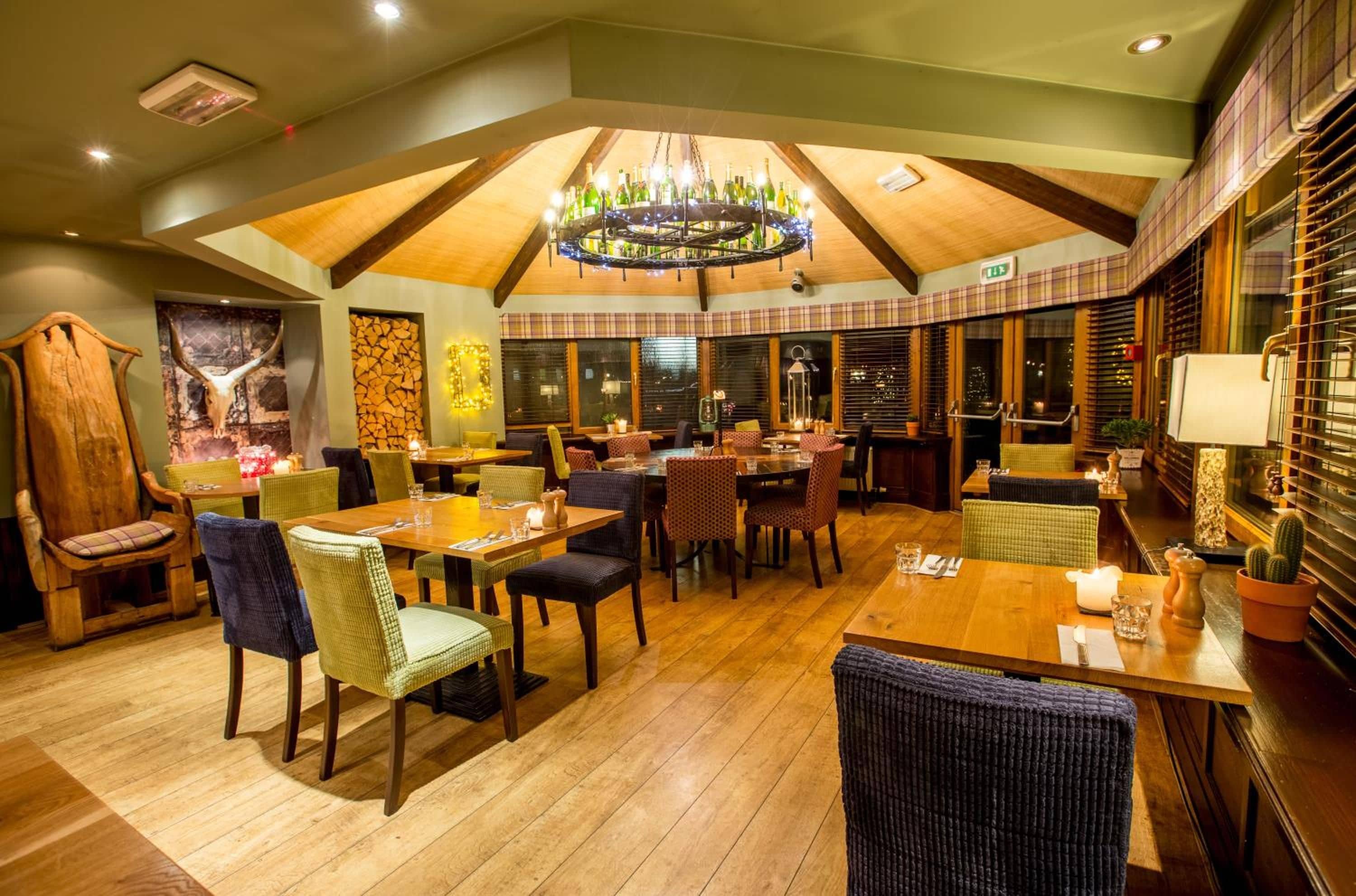 A restaurant with a high ceiling, wooden beams, and a chandelier. Tables and chairs are arranged for dining.