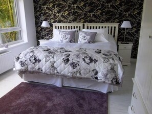 A bedroom with a floral bedspread, two bedside lamps, a white headboard, and a window with a view of greenery. - The Cock and Bull (Aberdeen)