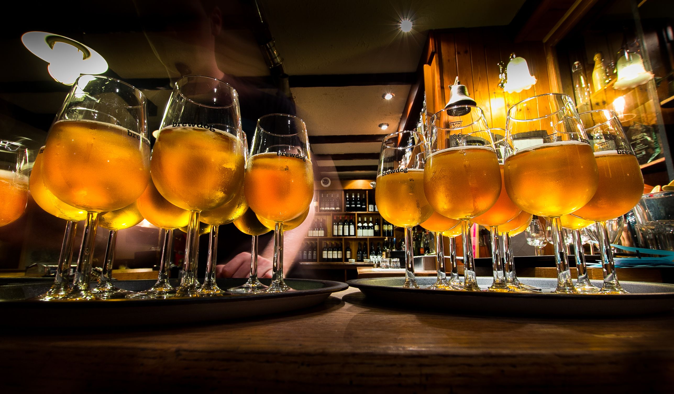 Um bar com vários copos de cerveja em bandejas, uma pessoa ao fundo e prateleiras com garrafas.