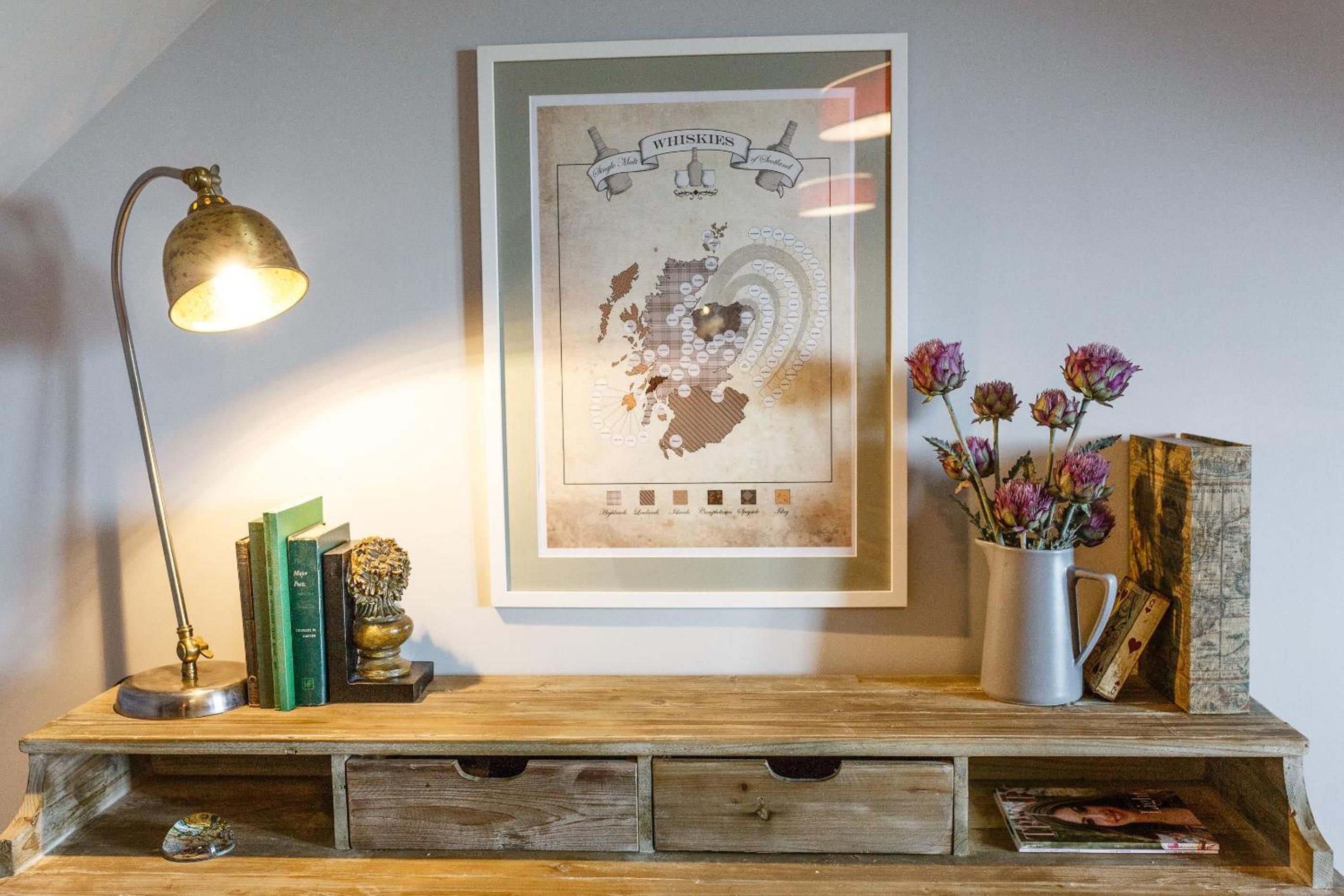A wooden desk with a framed map, a lamp, books, a vase with flowers, and a vintage-style lamp.