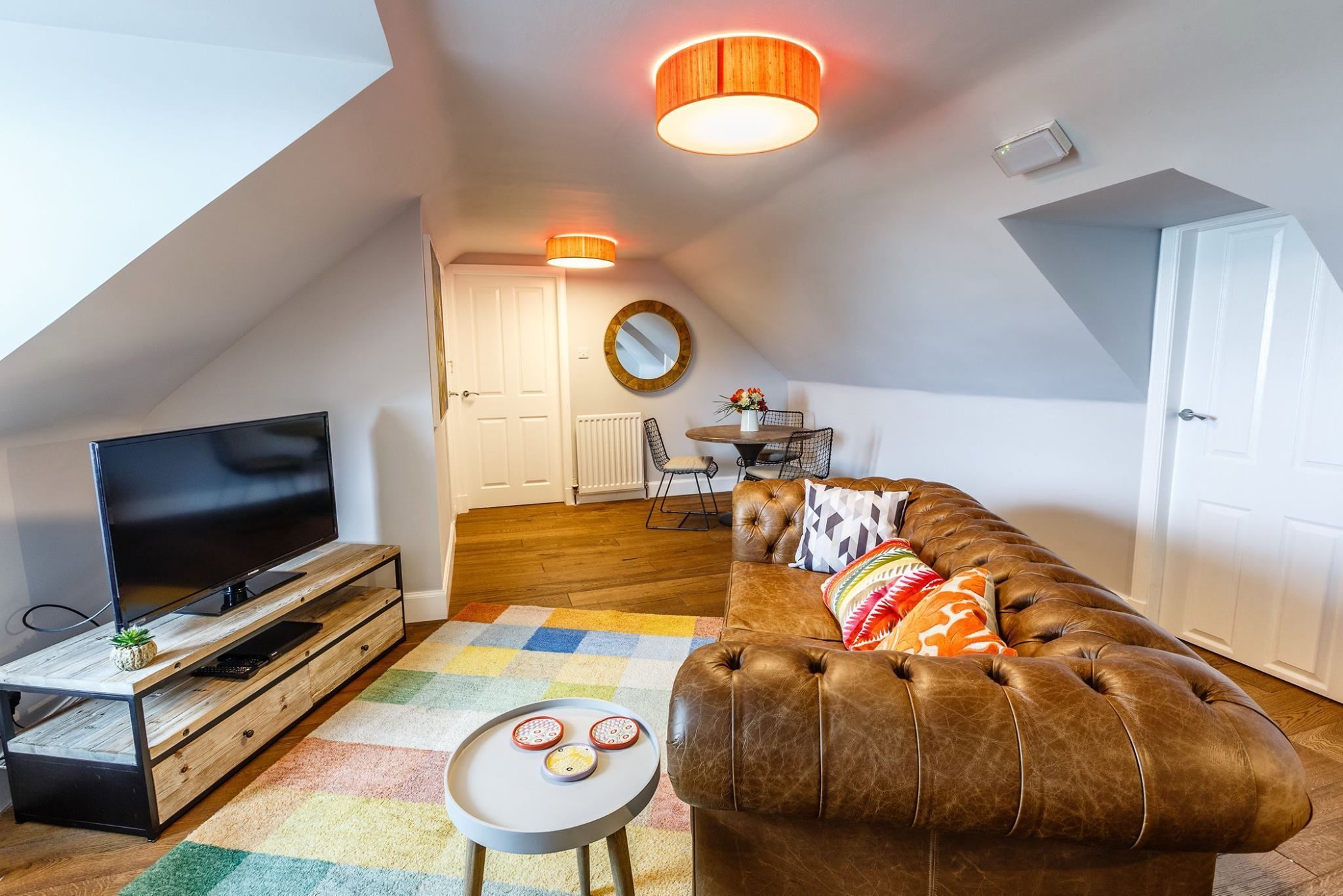 A cozy loft apartment with a large brown leather sofa, a flat-screen TV on a wooden stand, a colorful rug, and a small round table with decorative items.
