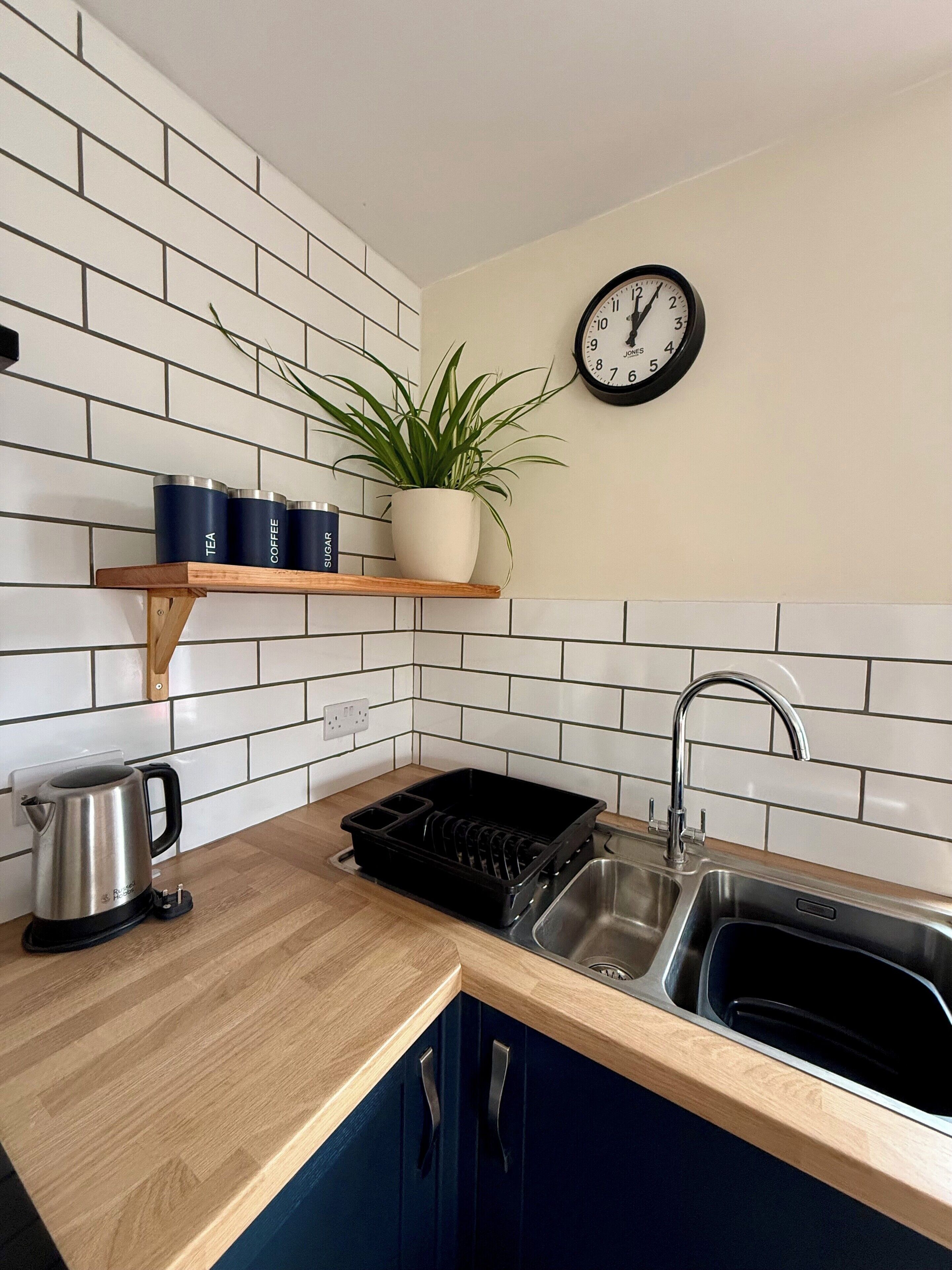 A kitchen with white tiled backsplash, a wooden countertop, a sink, a kettle, a plant, and a clock.
