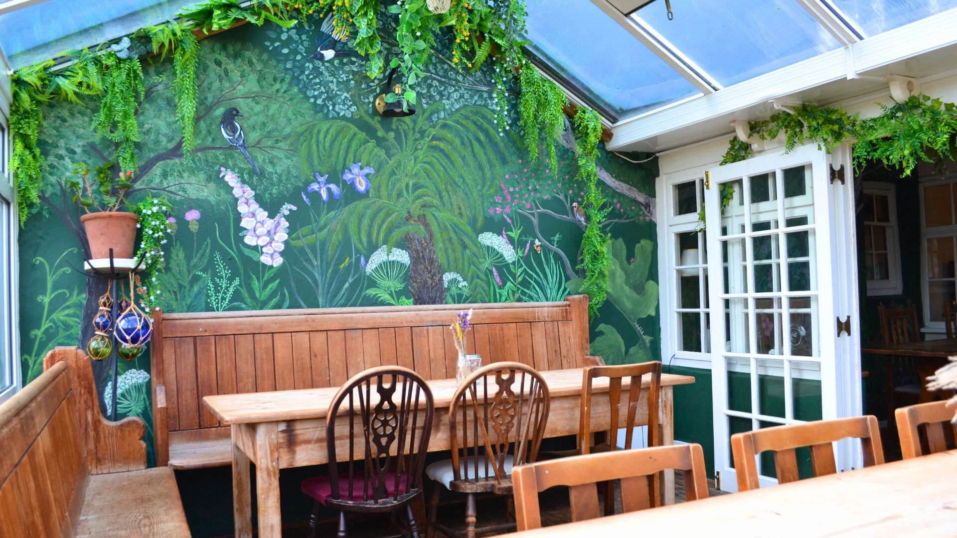 Un banc et une table en bois, placés devant une fresque murale verte représentant des oiseaux et du feuillage.