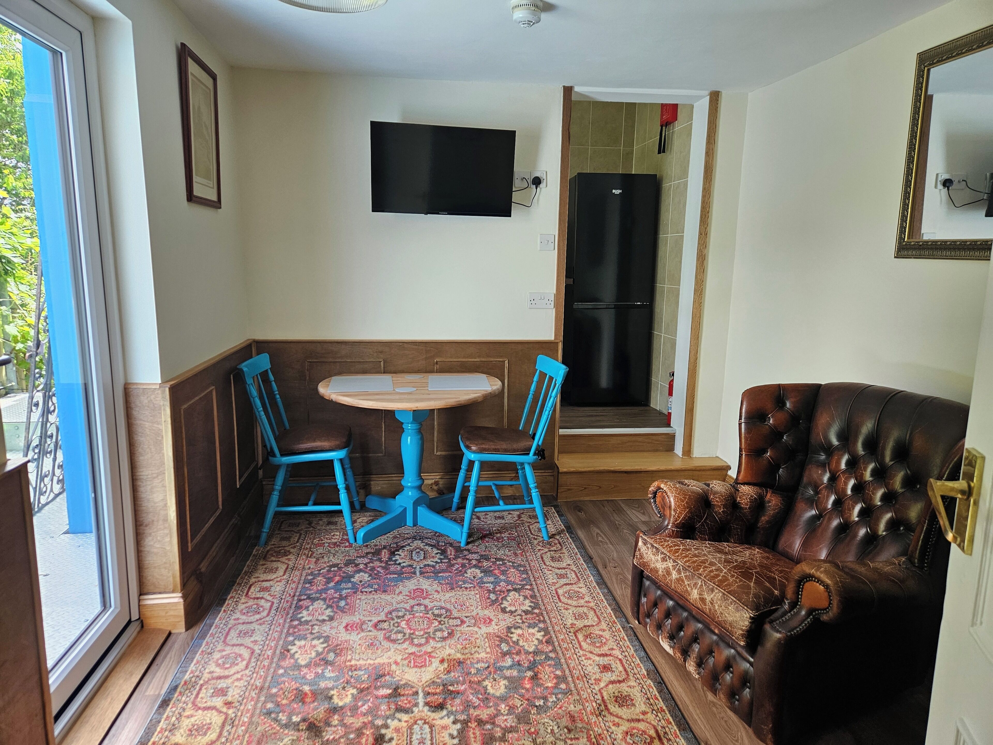 Una habitación con una mesa redonda, dos sillas azules, un sofá de cuero marrón, un tapete estampado, un televisor montado en la pared y un refrigerador al fondo.