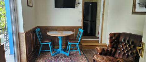 Una habitación con una mesa redonda, dos sillas azules, un sofá de cuero marrón, un tapete estampado, un televisor montado en la pared y un refrigerador al fondo.