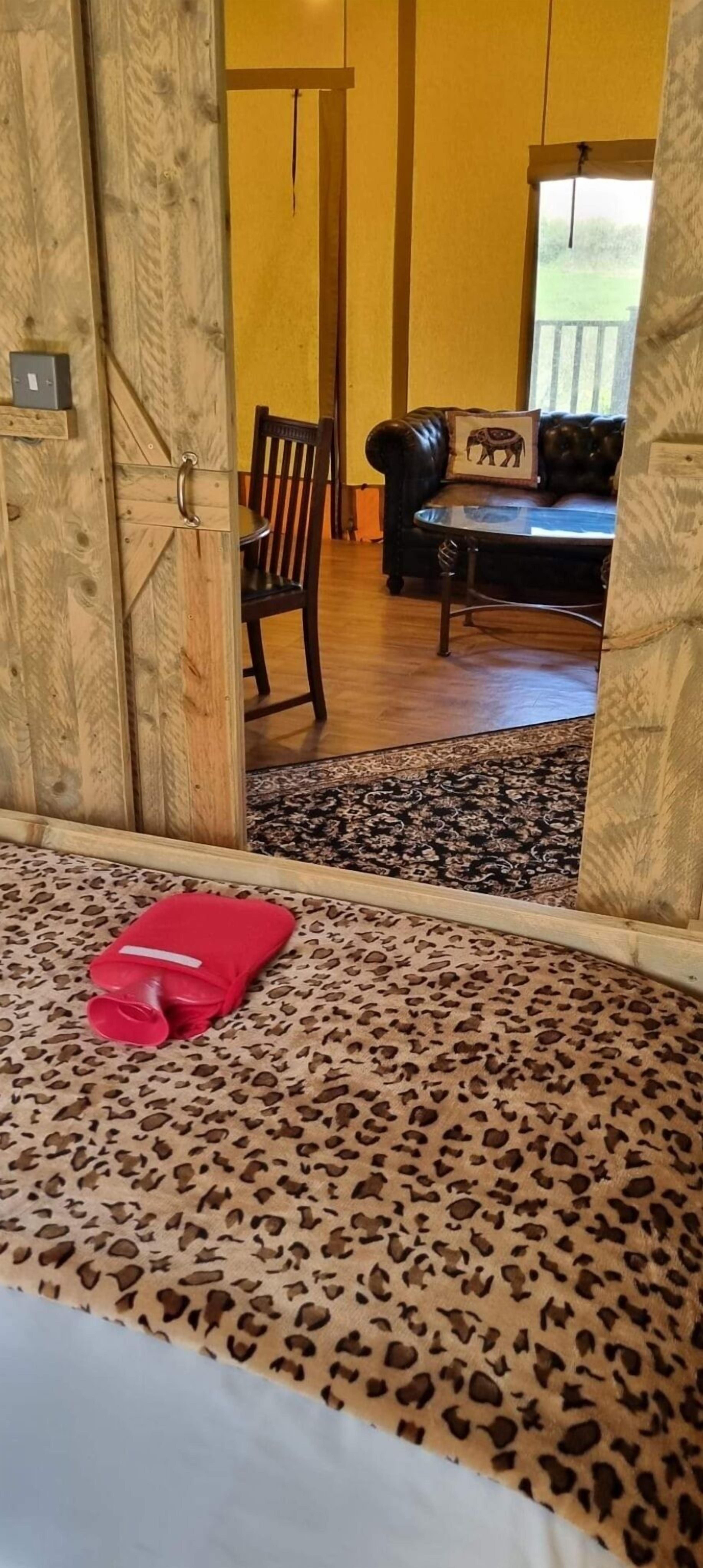 Quarto com carpete estampado de leopardo, almofada vermelha, porta de madeira e vista para uma sala de estar com sofá de couro e uma almofada.