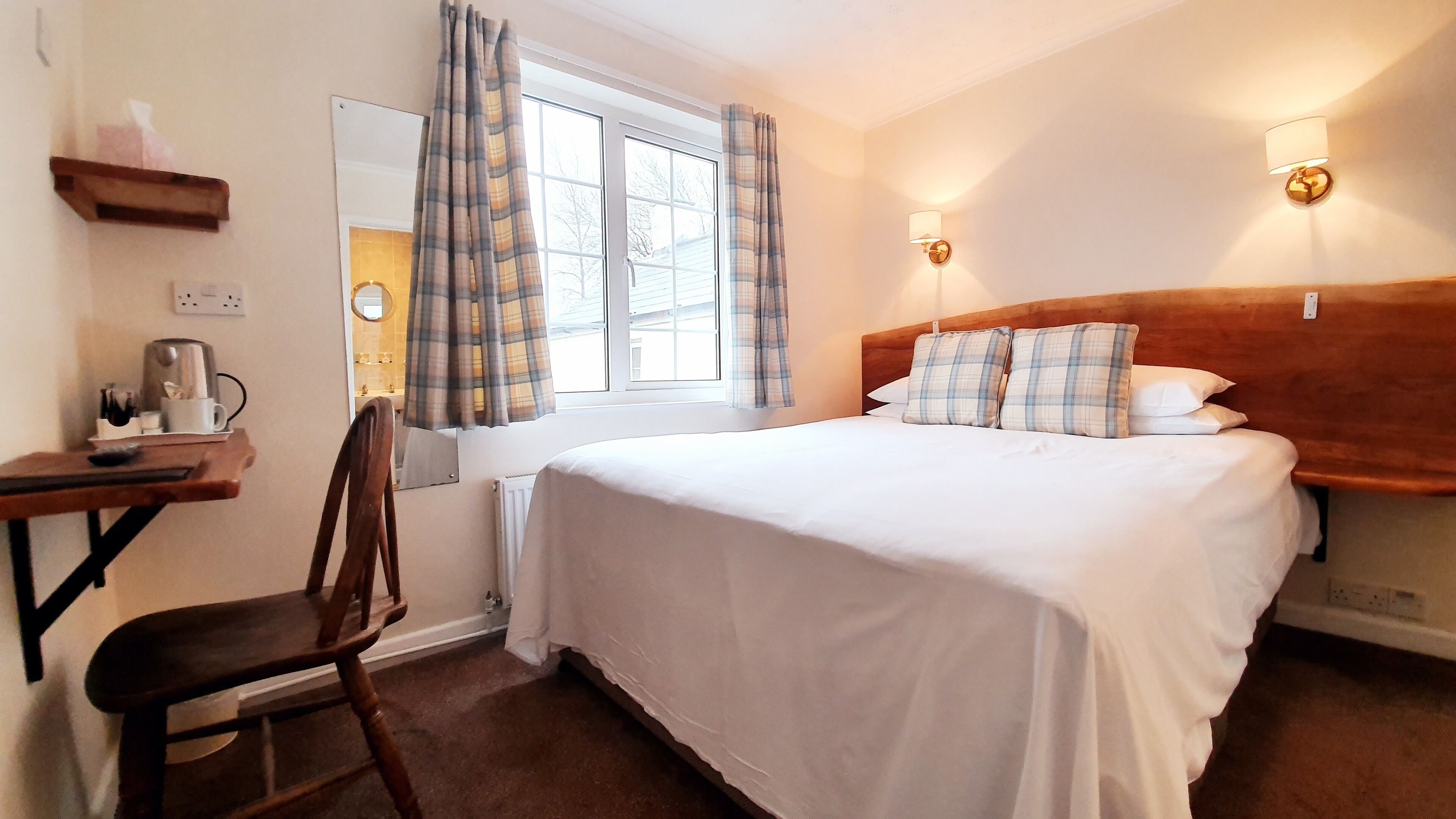A hotel room with a bed, a desk with a chair, a window with curtains, and a wall-mounted lamp.