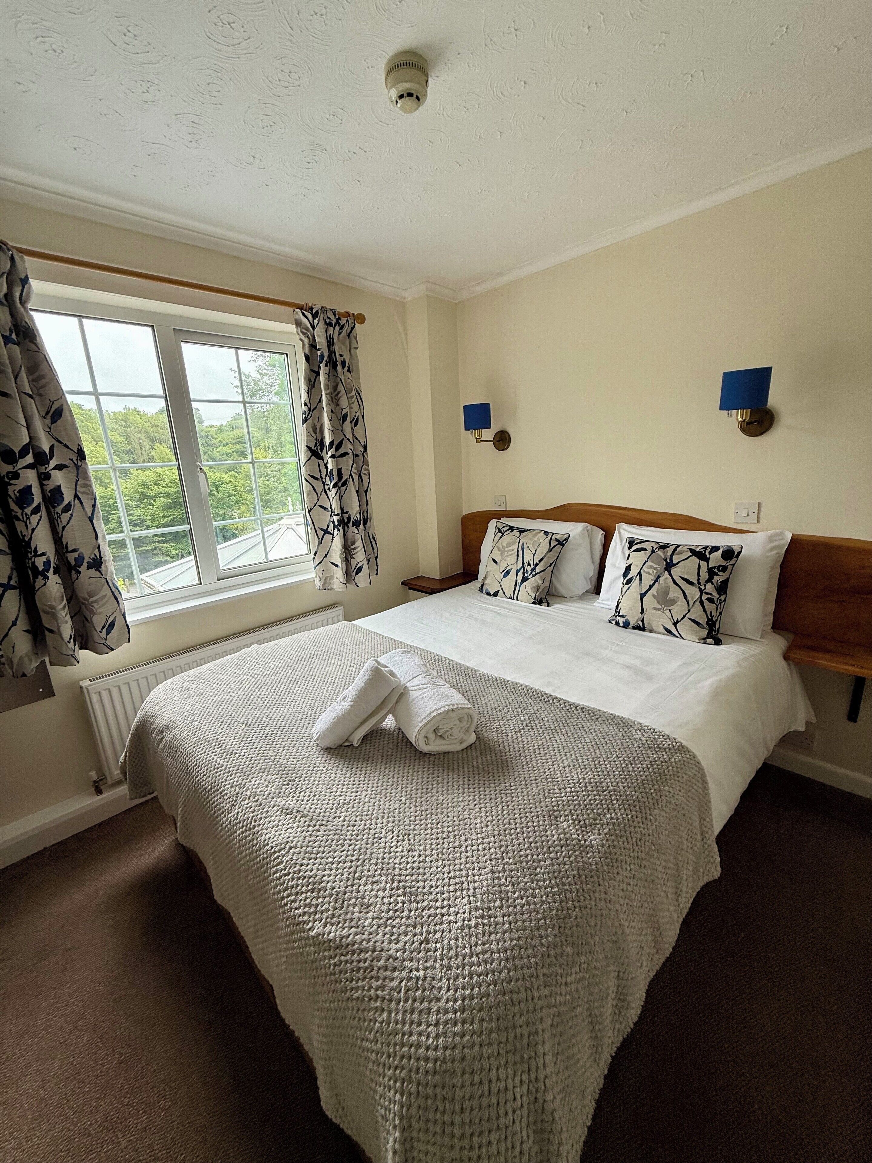 A hotel room with a large bed, two wall-mounted lamps, a window with a view of greenery, and a ceiling light.