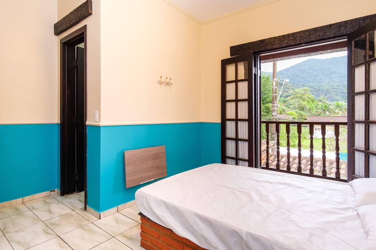 A room with a bed, a door, a window with a view of greenery and mountains, and a wall-mounted light fixture.