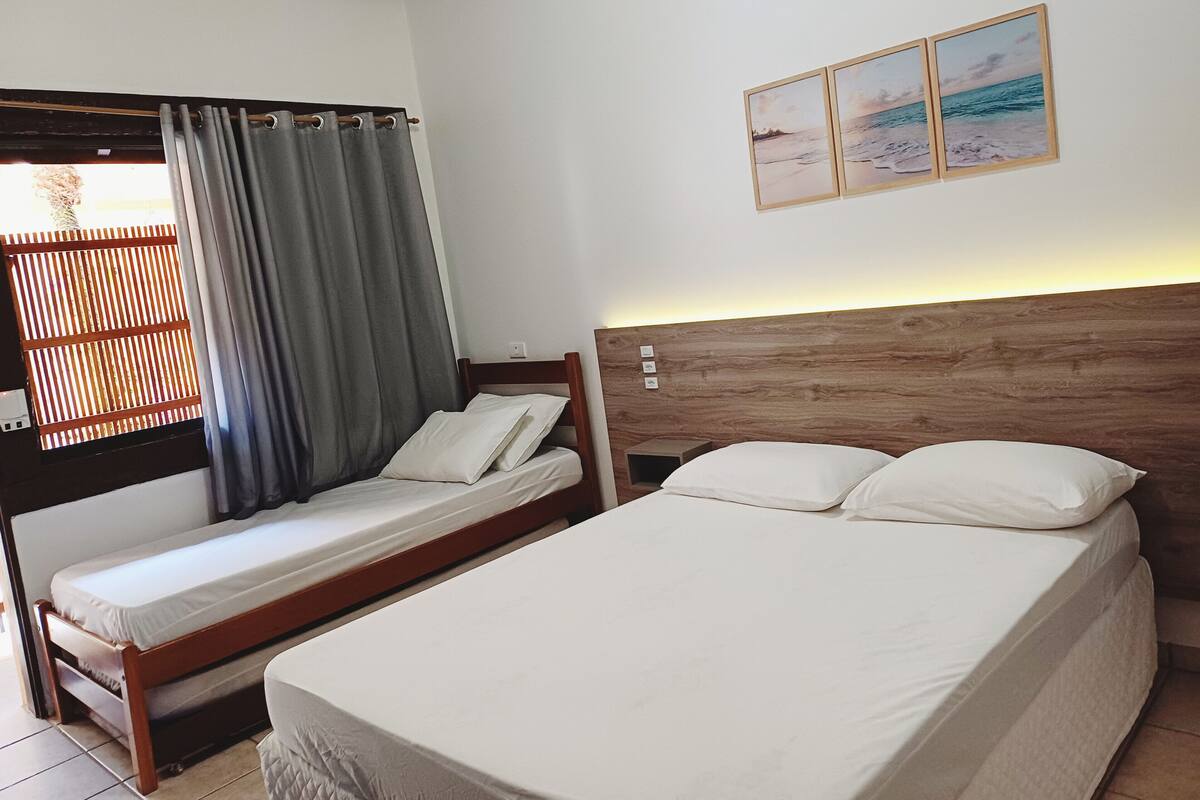 A room with two beds, one with a wooden headboard and the other with a plain headboard. There is a window with curtains, a wall-mounted picture, and a small shelf with items.