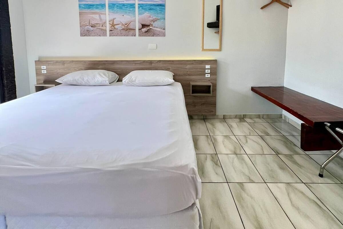 A hotel room with a bed, a wall-mounted air conditioner, a wooden headboard, a small shelf, and a mirror.