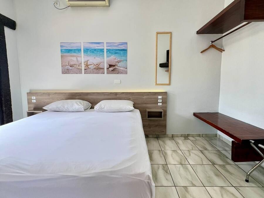 A hotel room with a bed, a wall-mounted air conditioner, a wooden headboard, a small shelf, and a mirror.