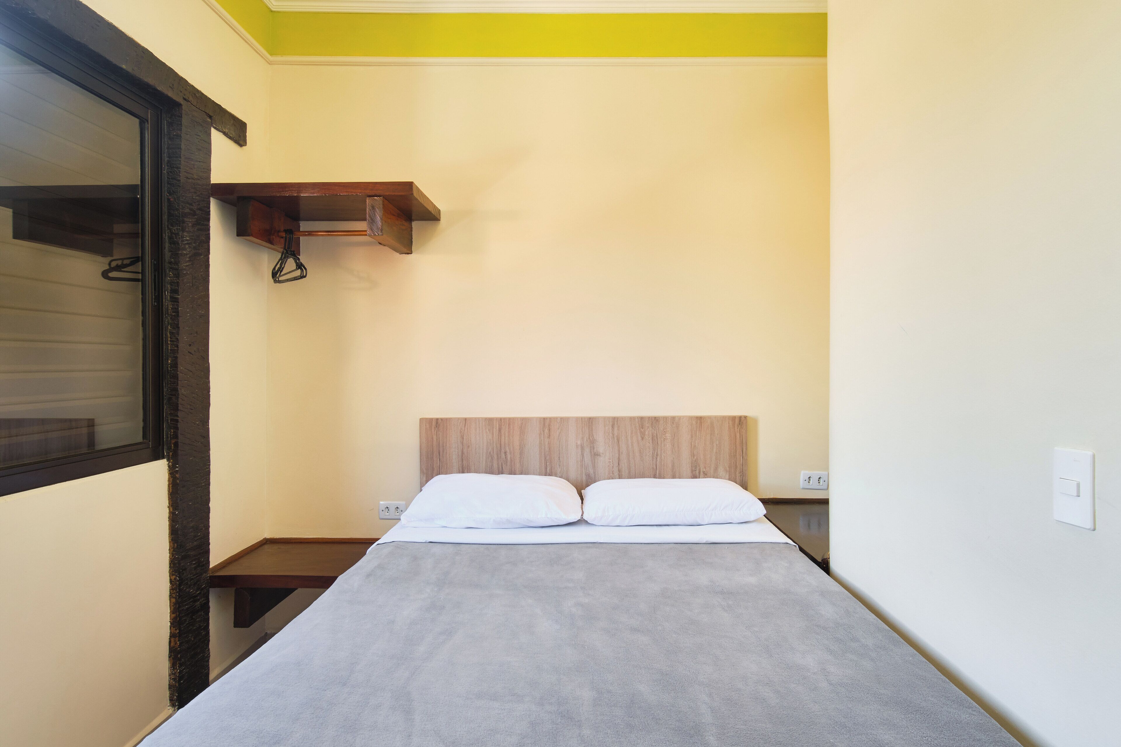 A small, narrow hotel room with a bed, a wooden headboard, a small shelf, and a window with blinds.