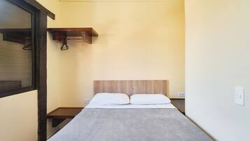 A small, narrow hotel room with a bed, a wooden headboard, a small shelf, and a window with blinds.