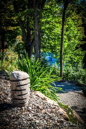 A stone structure surrounded by pebbles and green plants, with trees and a blue body of water in the background.
