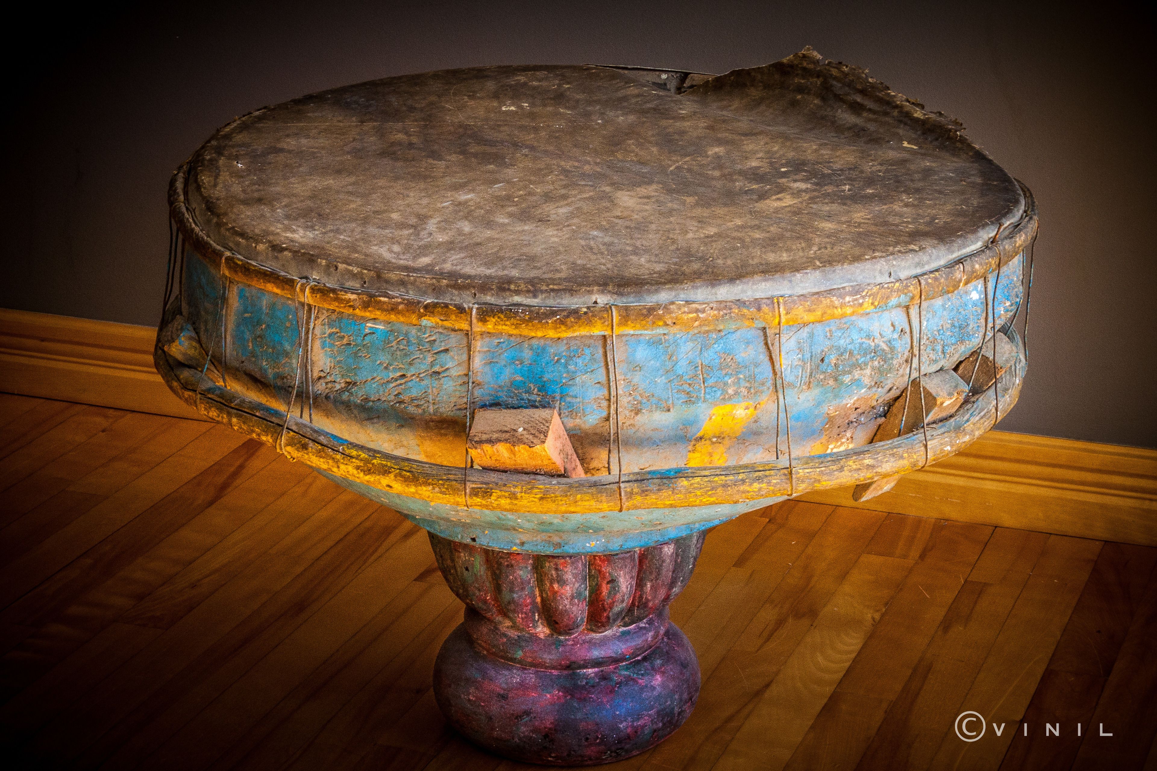 A drum with a worn, colorful exterior, placed on a wooden floor against a dark wall.