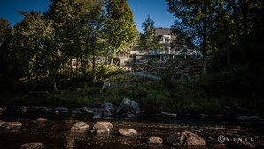 Vue sur une maison en pierre avec balcon, donnant sur la rivière, entourée d’une végétation dense et bordée d’un lit de rivière rocheux.