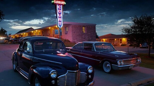 To klassiske biler parkeret foran en motel med et neonlys, der viser ledige værelser.