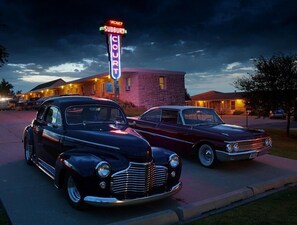 Dos autos antiguos estacionados frente a un motel con un letrero de neón que indica disponibilidad.