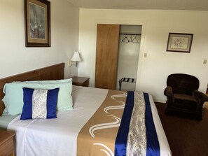 Ein Hotelzimmer mit einem ordentlich bezogenen Bett, einem Holfnachtkästchen, einem Sessel, einem Kleiderhaken und einer Tür zu einem Badezimmer.